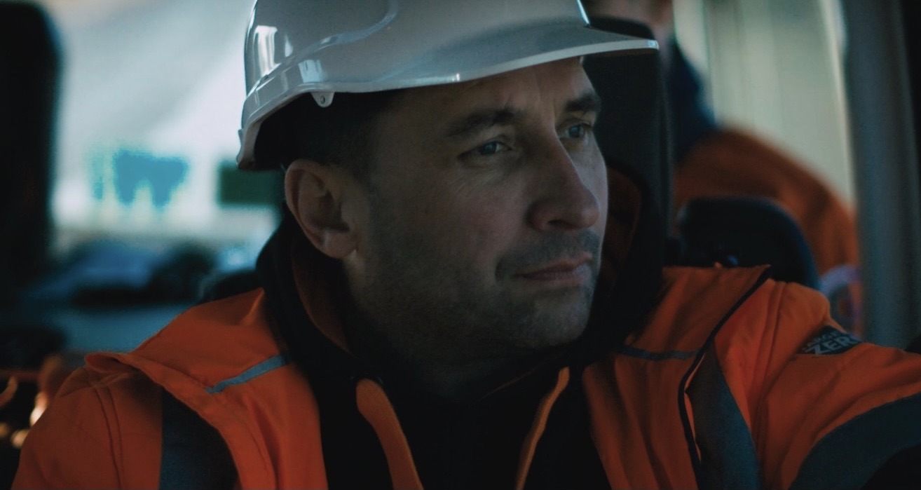 A line worker in a white hard hat and orange hi-vis jacket sitting in a crew vehicle cab
