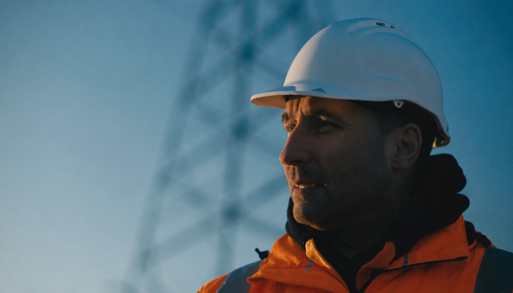 A line worker in a white hard hat and orange hi-vis jacket in side profile, with a transmission tower behind him