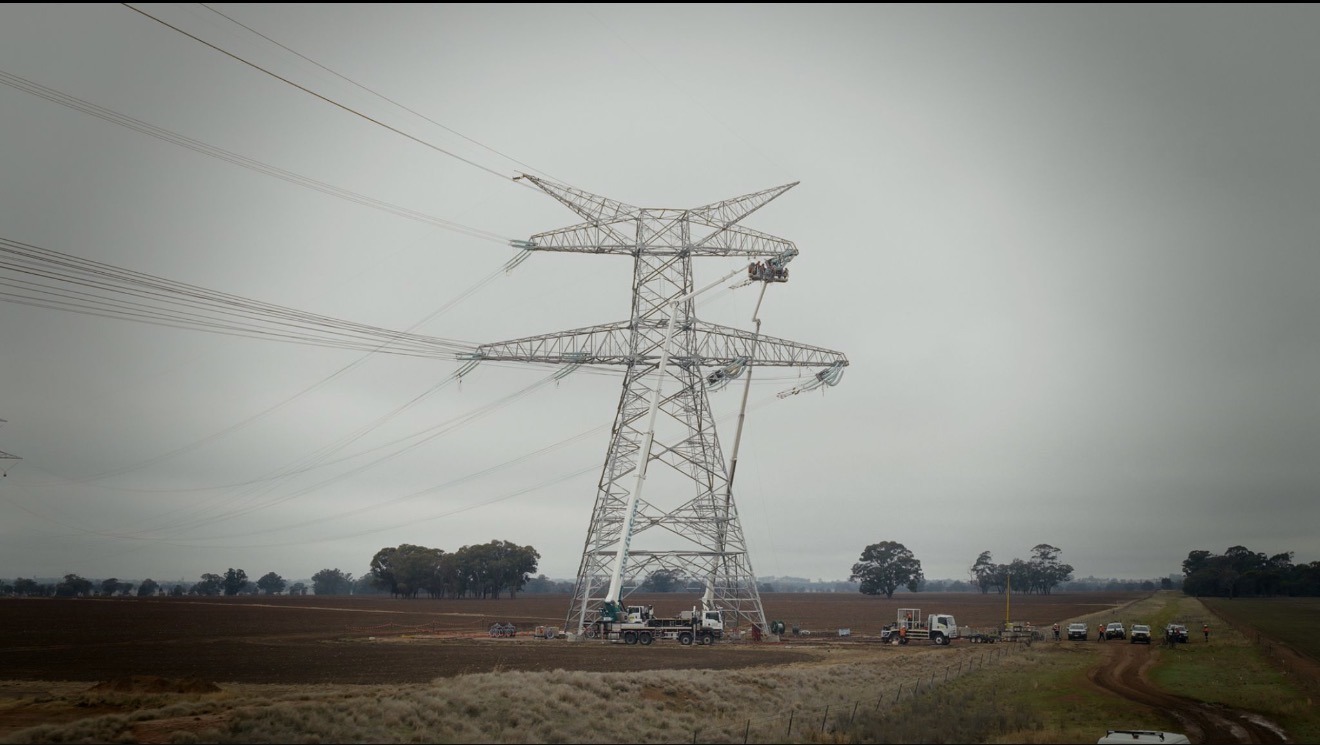 A transmission tower under construction with a crane, crew vehicles and workers on a rural site